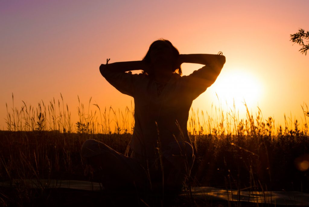 Silueta de una persona sentada en postura de meditación con manos detrás de la cabeza al amanecer en un campo