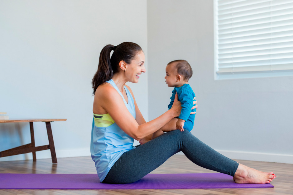 Mujer sentada en esterilla de yoga sosteniendo a su bebé mientras sonríen en una práctica de yoga postnatal