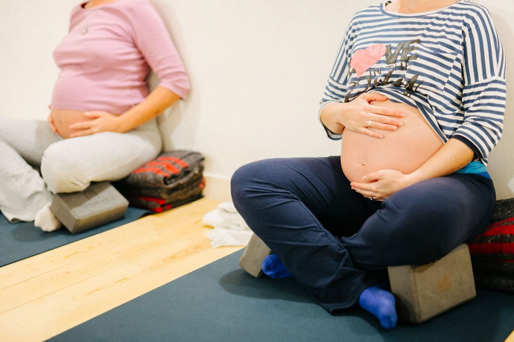 Dos mujeres embarazadas sentadas en bloques de yoga sobre esterillas, con las manos apoyadas en el vientre en una postura de relajación