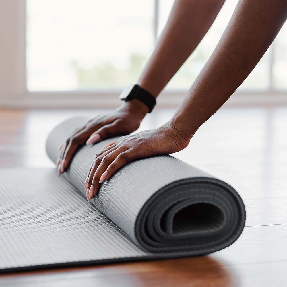 Manos femeninas con reloj inteligente enrollando una esterilla de yoga gris sobre suelo de madera